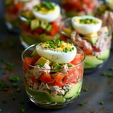 Bunte Schichtsalate in Gläsern mit Tomaten, Avocado, Thunfisch und gekochtem Ei on top. Mit Schnittlauch dekoriert.